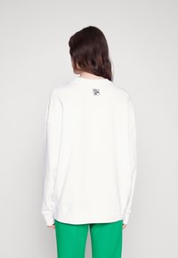 Sudadera blanca de corte relajado, con un logo bordado "HUGO" en negro en la parte posterior y puños acanalados. Combinada con pantalones verdes.