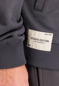 Mörkgrå sweatshirt med ribbade muddar och en vävd etikett. Etiketten visar "STUDIO EDITION" och "ALPHA INDUSTRIES INC." i svart text.