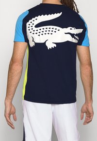 Krátký rukáv sportovního trička tmavě modré barvy s velkým bílým krokodýlím logem na zádech a modrými a neonově žlutými akcenty na rukávech.