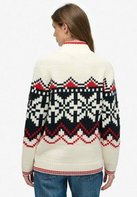 Pull en tricot blanc avec des motifs géométriques noirs, rouges et crème. Il est doté d'un col haut et de poignets et d'un ourlet côtelés.