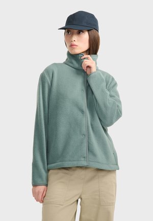 Veste en polaire vert clair avec un col haut et des boutons-pression, associé à une casquette bleu marine. Le tissu a une apparence douce et texturée.