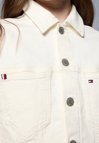 Krémová džínová bunda se stříbrnými knoflíky, skládacím límcem a kontrastními kapsami zdobenými červeným, bílým a modrým prošitím.