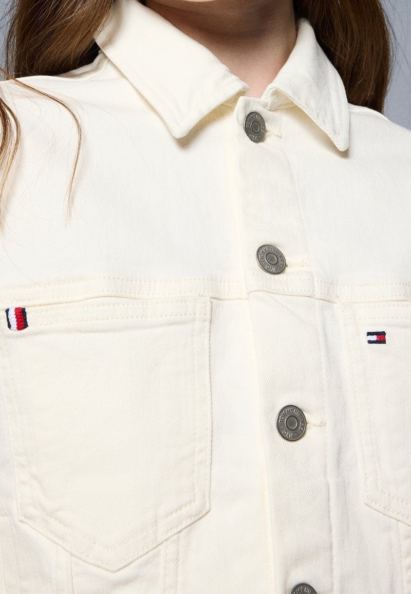 Krémová džínová bunda se stříbrnými knoflíky, skládacím límcem a kontrastními kapsami zdobenými červeným, bílým a modrým prošitím.