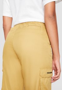 Osoba ubrana w musztardowe spodnie cargo z logo "CONVERSE" na bocznej kieszeni oraz w białą koszulę z krótkim rękawem, widoczna z tyłu.
