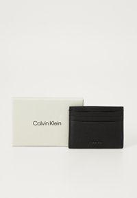 Svart skinn kortholder har flere spor, en teksturert overflate og et preget "Calvin Klein"-logo. Presenteres i en hvit eske.