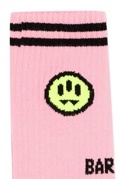 Chaussette rose avec deux rayures noires près du haut, présentant un visage jaune-vert à trois yeux avec une moue et un texte noir partiellement visible.