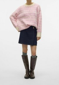 Femme portant un pull rose et crème avec le texte « PARIS », une jupe en denim foncé, des chaussettes grises et des bottes hautes marron foncé sur un fond blanc.