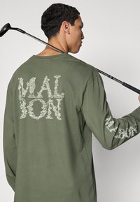 Μακρυμάνικο πουκάμισο ελιάς με λευκό κείμενο «MALBON» σε σχέδιο με λουλούδια στην πλάτη και στο δεξί μανίκι.