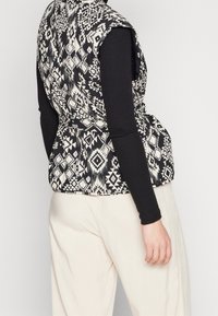 Svartvit mönstrad väst med peplum-design och utan ärmar, kombinerad med en svart långärmad topp och krämfärgade byxor.