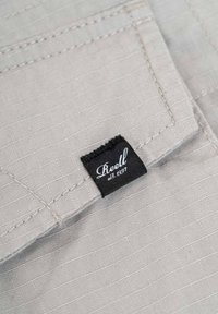 Hellgrauer Stoff mit strukturiertem Gewebe, ausgestattet mit einem auffälligen schwarzen Etikett mit weißer, geschwungener Schrift, auf dem "Reell est. 1997." steht.