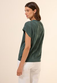 Knitted green sweater with a textured, patterned design, short sleeves, and a crew neckline, paired with white pants.