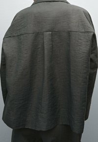 Veste oversize gris foncé en tissu texturé. Elle présente un col de chemise, des épaules tombantes et un détail plissé dans le dos.