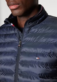 Námořnická pufovaná bunda s vysokým límcem, s horizontálně prošívaným designem, zipovým uzávěrem a malým logem s červenými, bílými a modrými akcenty.