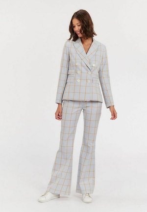 Femme portant un blazer croisé à carreaux bleu clair et beige ainsi qu'un pantalon évasé assorti avec des baskets blanches, souriante et regardant vers le bas.