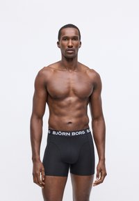 En mann uten skjorte står med ansiktet vendt fremover, iført svarte Björn Borg boxershorts mot en ensfarget hvit bakgrunn.