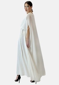 Robe blanche fluide en tissu léger, avec des manches façon cape et une taille cintrée. Portée avec des chaussures à talons noires.