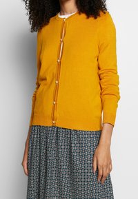 Gul stickad cardigan med rund halsringning, långa ärmar och framknäppning med pärlliknande knappar. Matchas med mönstrad kjol.