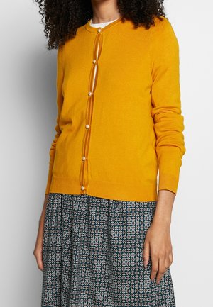 Geel gebreide cardigan met ronde halslijn, lange mouwen en voorste knoopsluiting met parelachtige knoppen. Gepaard met een gestreepte rok.