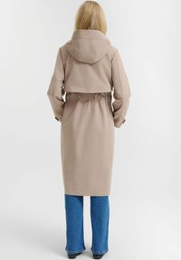 Tanfarbene, kapuzenbelegte Trenchcoat mit taillierter Silhouette und Rückenrüsche, kombiniert mit blauen Schlagjeans. Einheitliche Farbe, klare Linien und minimales Zubehör.