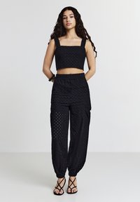 Donna con capelli lunghi e ricci, che indossa un top corto senza maniche nero e pantaloni in tessuto traforato coordinati, con polsini elastici e tacchi neri a strappo.