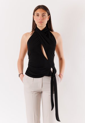 Donna con top nero senza maniche a fascia con scollo halter, apertura frontale e dettaglio annodato sul lato, abbinato a pantaloni beige a vita alta.