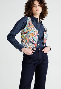 Young person with curly hair wearing a colorful patterned vest over a dark denim buttoned shirt and dark high-waisted pants.