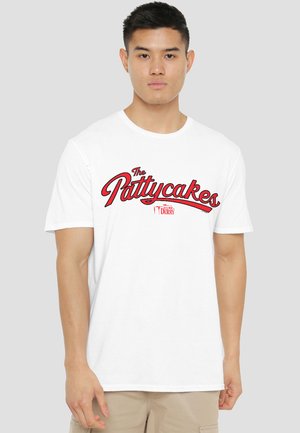 Young man wearing a white T-shirt with red text "The Pattycakes" and smaller text referencing "Derry" on a plain gray background.