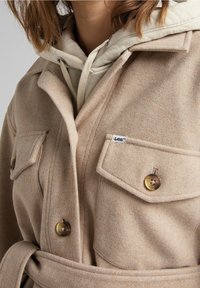 Primer plano de una persona que lleva un abrigo de lana beige con grandes botones, bolsillos de solapa, un cinturón y una sudadera con capucha beige claro debajo.