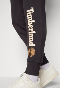 Svarta sweatshirtbyxor med "Timberland"-logotyp i beige på sidan och en träemblem. Kombinerat med vita sneakers och grå strumpor för kontrast.