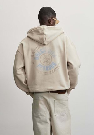 Mand iført beige hættetrøje med rundt Dolce Vita Pecador-logo på ryggen, beige bukser, solbriller, stående mod en ensfarvet lys baggrund.