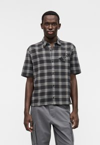 Πουκάμισο με κοντό μανίκι σε καρό μοτίβο με γκρι και μαύρους τόνους, με τσέπη στο μπροστινό μέρος που φέρει σχέδιο αστέρι. Το ύφασμα φαίνεται μαλακό και έχει άνετη γραμμή.