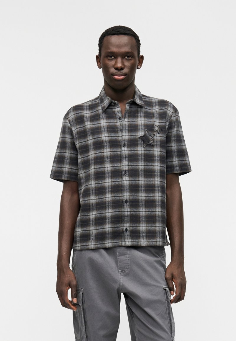 Πουκάμισο με κοντό μανίκι σε καρό μοτίβο με γκρι και μαύρους τόνους, με τσέπη στο μπροστινό μέρος που φέρει σχέδιο αστέρι. Το ύφασμα φαίνεται μαλακό και έχει άνετη γραμμή.