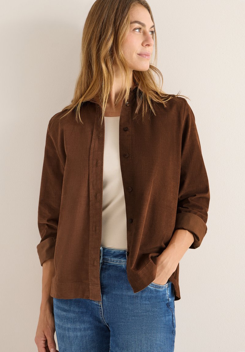 Camicia in corduroy marrone a maniche lunghe, con bottone frontale e orlo arrotondato. Abbinata a una maglietta crema e jeans in denim blu.