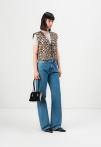 Vrouw in een panterprint vest over een grijs T-shirt, blauwe wijde jeans, zwarte puntige schoenen, en met een zwarte handtas.