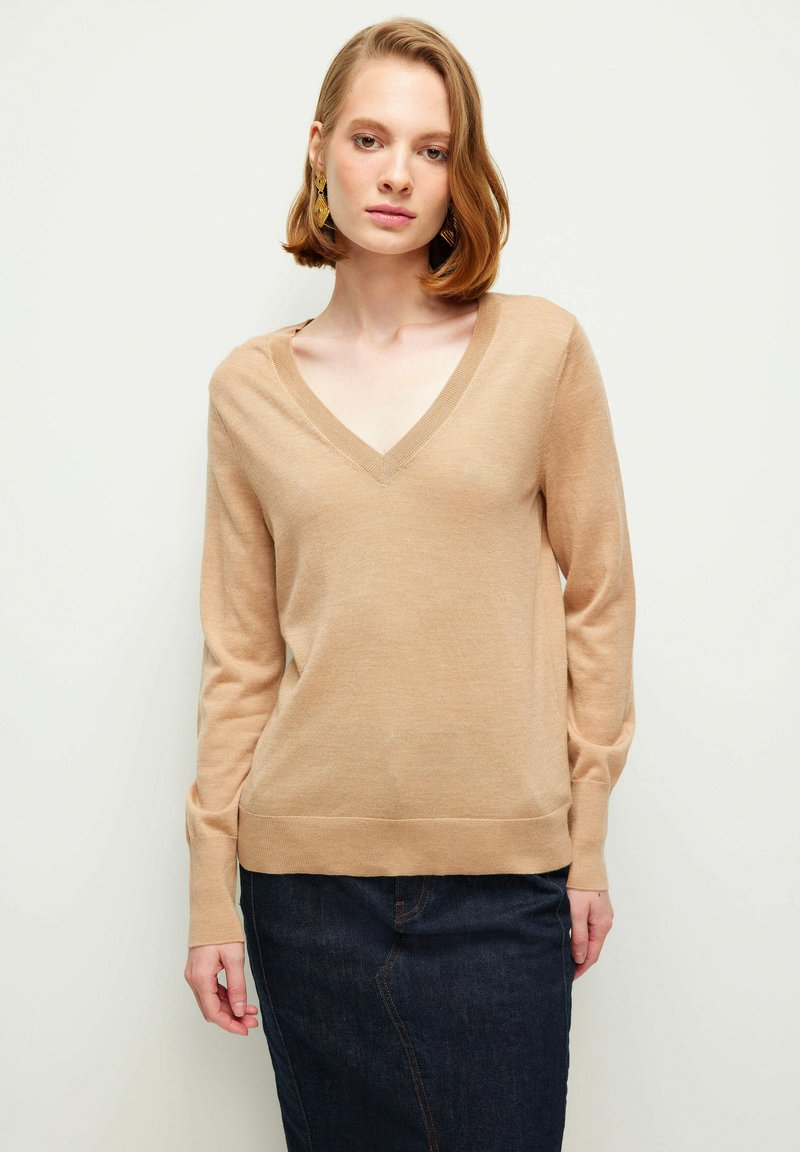 Hellbeige V-Ausschnitt-Pullover mit langen Ärmeln, weichem Stoff, gerippten Bündchen und Saum, kombiniert mit einem dunklen Jeansrock.