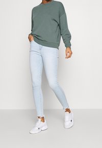 Sudadera verde grisácea con hombros caídos, combinada con jeans ajustados azul claro con dobladillos deshilachados y zapatillas blancas.