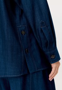 Zbliżenie ręki osoby obok mankietu i bocznych guzików ciemnoniebieskiej kurtki dżinsowej z brązowym przeszyciem.