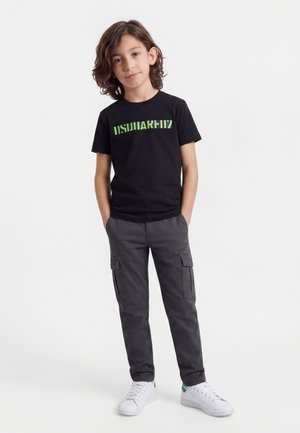 Jonge jongen met bruin haar staat met handen in de zakken, draagt een zwart T-shirt met print, grijze cargobroek en witte sneakers.