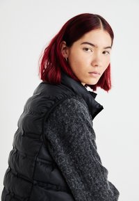 Zwarte puffer vest met een geborduurd patroon, gecombineerd met een donkergroen wollen trui. De vest heeft een opstaande kraag en een mouwloos ontwerp.