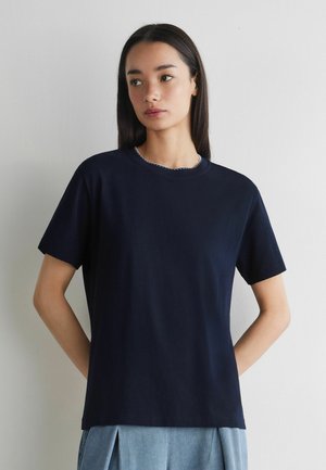 Ung kvinde med langt mørkt hår iført en simpel mørkeblå t-shirt med korte ærmer og lyse cowboybukser, stående mod en hvid væg.