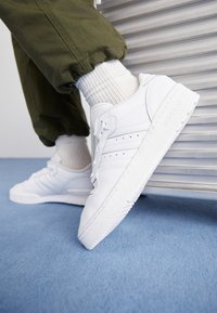 Vita sneakers med slät läder, perforerade detaljer och en texturerad sula, tillsammans med ribbade vita strumpor och olivgröna byxor.