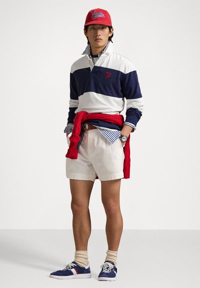 Marineblå og hvide stribede rugbytrøje med et rødt logo, cremefarvede shorts, rød sweater bundet om livet, blå sneakers og en rød kasket.