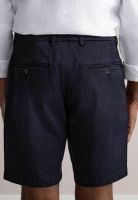 Navy-Shorts aus glattem Stoff, mit zwei Gesäßtaschen mit Knopfverschluss und einem sauberen Saum. Getragen mit einem weißen Hemd.