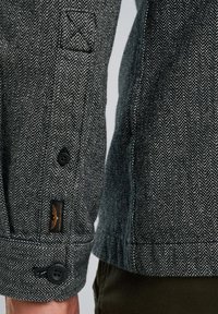 Sivi sako rukav s crnim dugmetom, koji ima logotip na zakrpi, kontrastno šivanje i vidljive detalje križnog šavova.