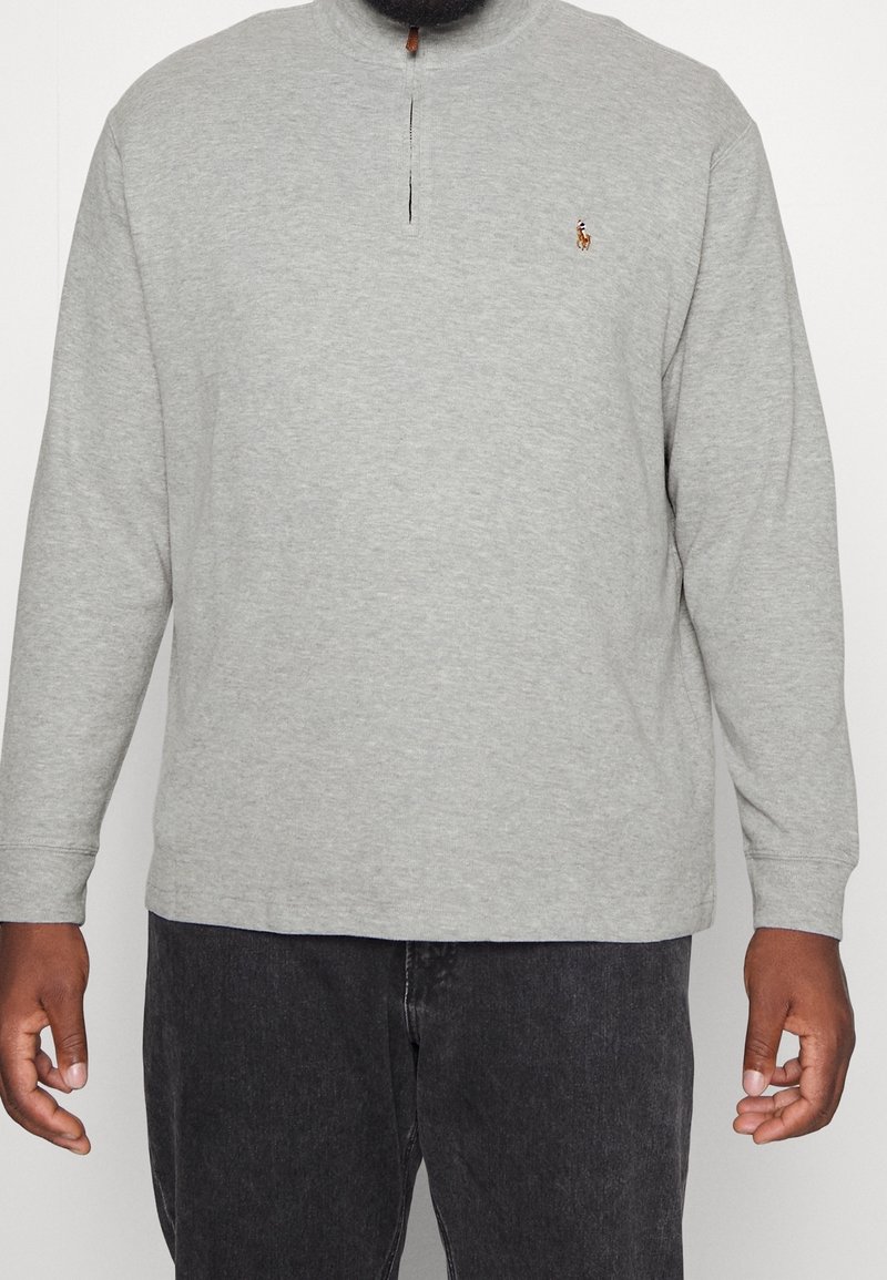 Man som bär en ljusgrå långärmad tröja med halv dragkedja och liten broderad logotyp, i kombination med svarta jeans.