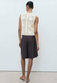 Cream crochet crop top with square patterns and a high neckline, paired with dark brown knee-length shorts. Simple footwear and minimal accessories.