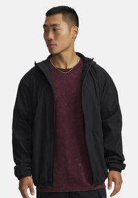 Young man looking left wearing a black zip-up hooded jacket over a maroon T-shirt and a thin gold chain necklace.