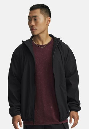 Young man looking left wearing a black zip-up hooded jacket over a maroon T-shirt and a thin gold chain necklace.