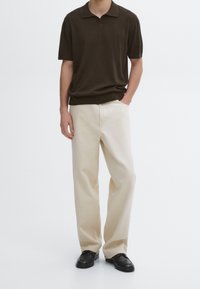 Homme portant un polo en maille à manches courtes marron foncé, un pantalon large crème, des mocassins en cuir noir, debout avec une main dans la poche sur un fond blanc.