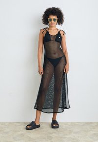 Črna mrežasta pokrivalna obleka z naramnicami za halter, prozorno teksturo in stranskimi razporki. Nosi se preko črnega bikini kompleta, v kombinaciji s črnimi natikači in zelenimi sončnimi očali.